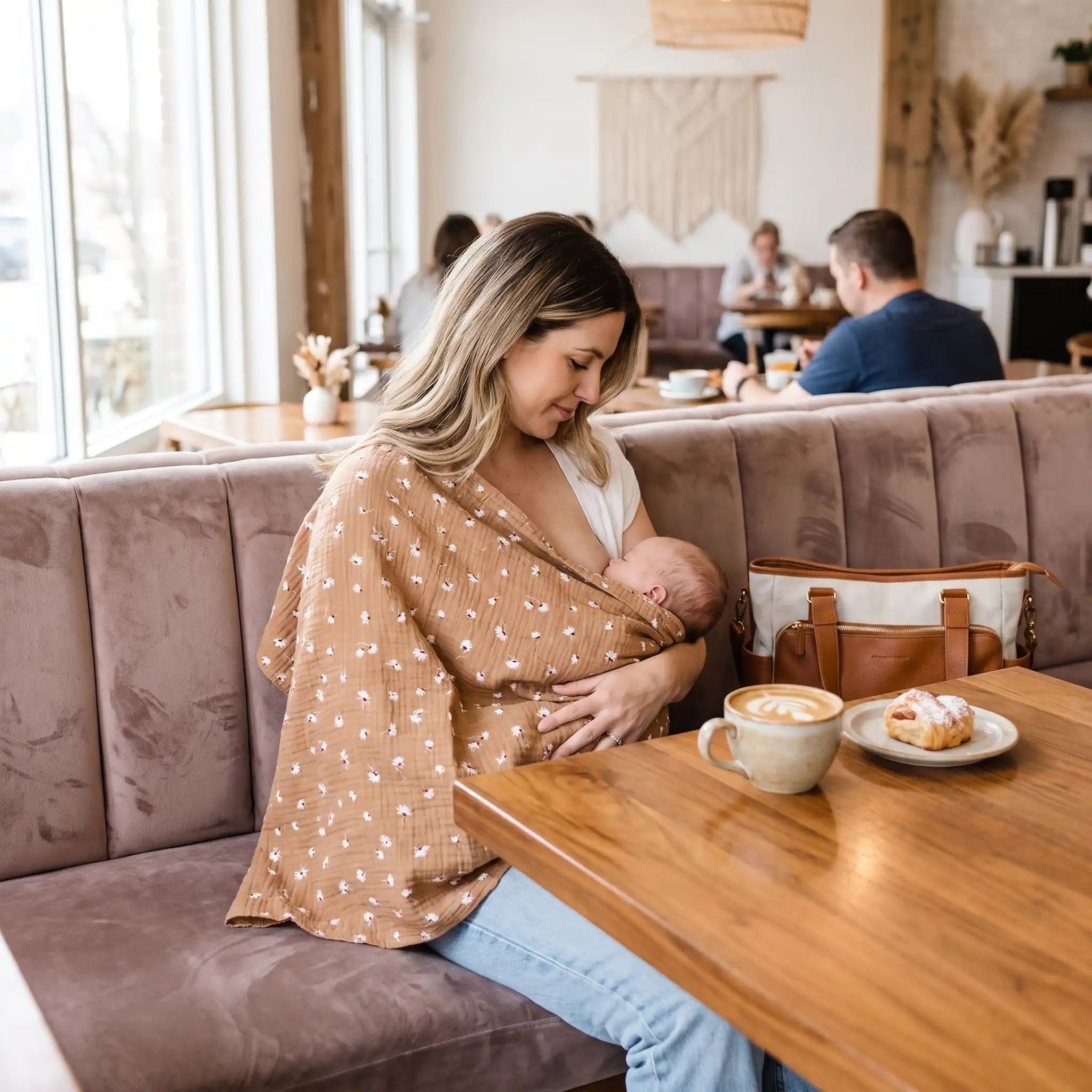 Cape d'allaitement beige rosé à petites fleurs portée par une maman allaitant son bébé sur une banquette de café.